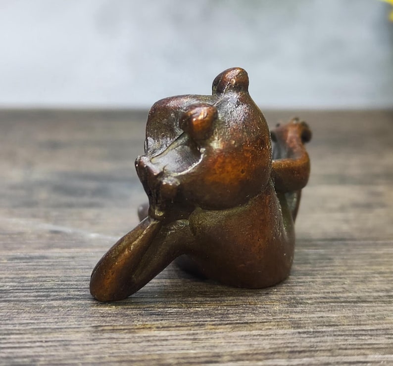 May include: A small, bronze-colored sculpture of a bear in a seated position. The bear is depicted with its arms raised and its head tilted back. The sculpture has a smooth, polished finish and appears to be made of metal.