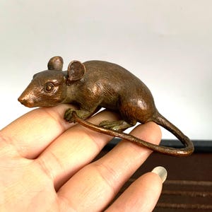 May include: A small bronze sculpture of a rat with a curled tail. The rat is depicted in a sitting position with its head turned to the side.