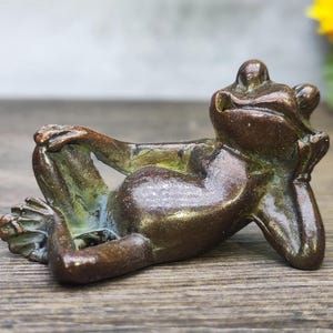 May include: A bronze frog sculpture in a relaxed pose, resting its head on its hand and crossing its legs. The frog has a weathered, aged appearance with hints of green and blue patina, suggesting an antique or vintage piece.