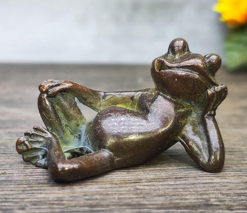 May include: A bronze frog sculpture in a relaxed pose, resting its head on its hand and crossing its legs. The frog has a weathered, aged appearance with hints of green and blue patina, suggesting an antique or vintage piece.