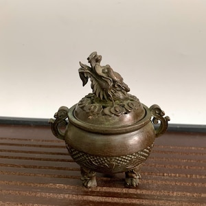以下が含まれることがあります： 蓋に龍の装飾が施されたブロンズ製のお香立て。丸いお香立てには、詳細な花の模様と3つの小さな足があります。蓋の上には龍の彫刻があります。お香立ては濃いブロンズ色です。