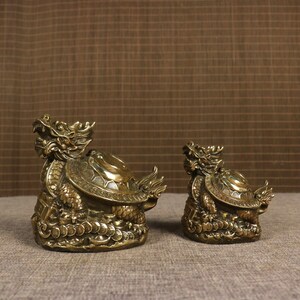 May include: Two bronze-colored dragon turtle figurines. The dragon turtles have detailed scales, a dragon head, and a turtle shell. The figurines are on a textured surface with a brown background. The dragon turtles are a symbol of longevity and prosperity.