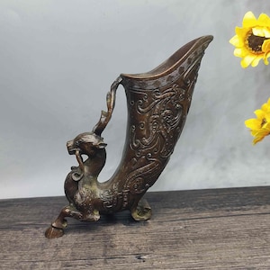 Op de afbeelding: Bronskleurig, sierlijk vat met een hertachtige figuur aan de zijkant. Het vat heeft een gebogen, hoornachtige vorm met gedetailleerde reliëfpatronen. Het hert heeft een gewei en staat in een rennende pose.