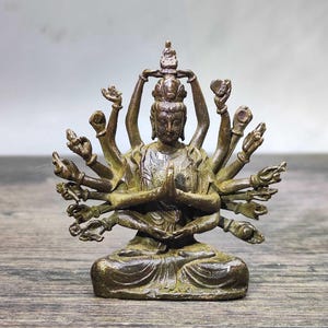 Può includere: Statua in bronzo di una divinità seduta con più braccia, mani in preghiera e un'espressione serena. La figura è adornata con dettagli intricati e si siede in una posa meditativa. La statua è su una superficie di legno.