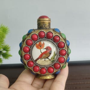 May include: Ornate, antique-style bottle with a bird and flower design. The bottle features a central image of a red bird and orange flower, surrounded by red, blue, and green decorative stones. The bottle has a gold-colored metal base and cap.