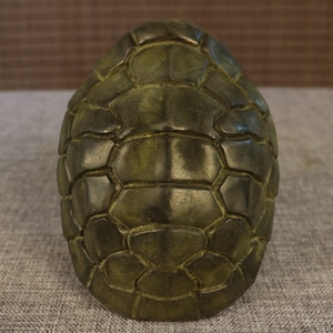 May include: A detailed, olive green turtle shell sculpture. The shell is segmented with a textured, geometric pattern. The sculpture has a smooth, polished finish, and the color varies from dark to light green.