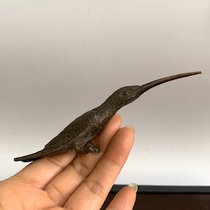 Pode incluir: Uma escultura de beija-flor em bronze com um bico longo e fino. A ave é retratada em perfil, com as asas dobradas e o corpo ligeiramente inclinado. A escultura tem uma textura detalhada e está sobre uma mão.