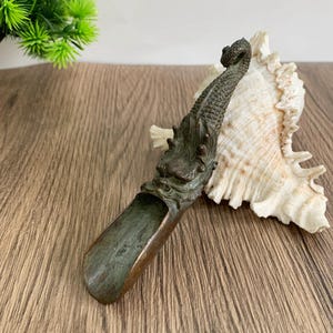 Puede incluir: Una cuchara de té de color bronce con un diseño de dragón. La cuchara tiene un cuenco largo en forma de artesa y una cabeza y cola de dragón detalladas. Se coloca sobre una superficie de madera junto a una concha marina.