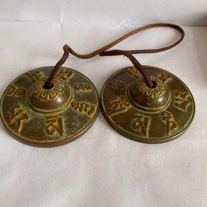 May include: Two antique bronze Tibetan singing bowls with embossed symbols and a leather cord. The bowls are circular with a central raised area and are connected by a brown leather cord. The bronze has a patina, suggesting age and use.