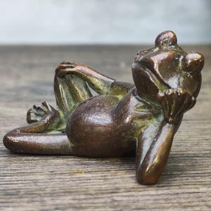 May include: A bronze frog figurine in a relaxed pose, resting on its elbows and knees. The frog's body is a dark brown color with hints of green, and it appears to be made of metal. The frog is lying on a wooden surface.