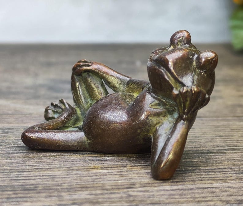 May include: A bronze frog figurine in a relaxed pose, resting on its elbows and knees. The frog's body is a dark brown color with hints of green, and it appears to be made of metal. The frog is lying on a wooden surface.