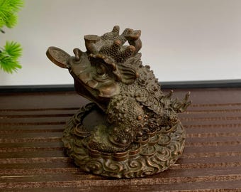 May include: A bronze-colored frog statue with a coin in its mouth and coins on its back. The frog sits on a base with a swirling design. The statue has a textured surface and is likely made of metal.