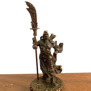 以下が含まれることがあります： 装飾的な刃が付いた槍を持った戦士のブロンズ像。戦士はゆったりとしたローブを着ており、あごひげを生やしています。像は、複雑なディテールが施された円形の台座の上に立っています。