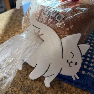 May include: A white cat-shaped bread bag clip. The clip is made of plastic and has a hook at the top. The cat has a smiling face and is holding a clear plastic bag. The clip is designed to keep bread fresh.