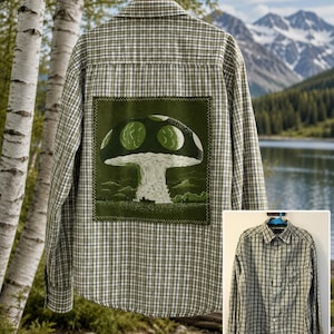 May include: A long-sleeve button-down shirt with a green and white plaid pattern. The back features a large, square, olive green patch with a graphic illustration of a mushroom, a small boat, and clouds, stitched onto the fabric with visible thread.