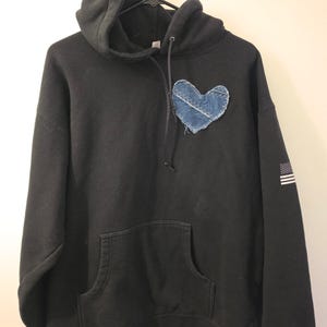 May include: A black hooded sweatshirt featuring a blue denim heart patch stitched onto the left chest area. The hoodie includes a front kangaroo pocket, long sleeves with a small white American flag graphic on the left sleeve, and black drawstrings.