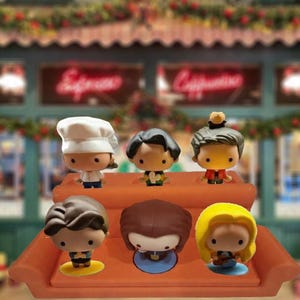 May include: Six cartoon character figurines are arranged on an orange platform. The figurines have varied hairstyles and outfits, including one with a chef's hat. The background shows a cafe with festive decorations.