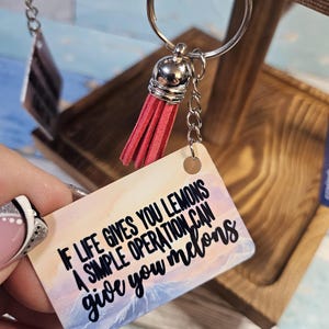 Puede incluir: Un llavero con un borla roja y una etiqueta rectangular que dice "If life gives you lemons a simple operation can give you melons". La etiqueta tiene un fondo de acuarela con una puesta de sol y montañas.
