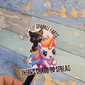 Puede incluir: Una pegatina con un gato negro montando un unicornio arcoíris. El texto dice "Giddy Up Sparkle Farts There's Chaos To Spread".