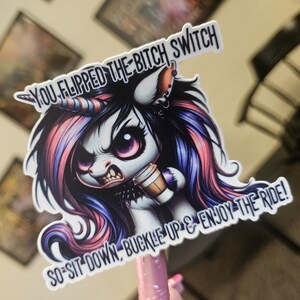 May include: A sticker featuring an illustration of an angry unicorn with pink, purple, and blue hair, holding a coffee cup. The text on the sticker reads "YOU FLIPPED THE BITCH SWITCH, SO-SIT DOWN, BUCKLE UP & ENJOY THE RIDE!"