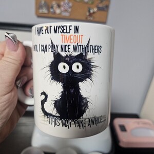 May include: White ceramic mug with a black cat illustration and the text "I have put myself in timeout until I can play nice with others. This may take a while..."