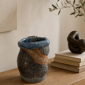 May include: A ceramic vase with an irregular, organic shape featuring a textured dark gray body with a lace-like pattern, a smooth tan diagonal band, and a thick, glossy blue rim. The base of the vase is accented with a light blue painted design.