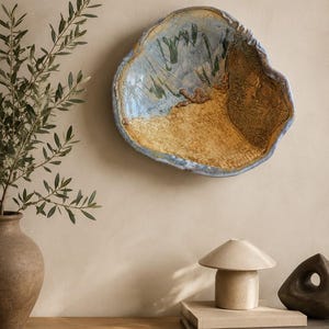 Może przedstawiać: Nieregularna, ręcznie formowana ceramiczna misa ścienna o teksturowanej powierzchni z mieszanką niebieskich, beżowych i brązowych szkliw. Wnętrze posiada wzór przypominający krajobraz z wypukłymi elementami glinianymi oraz falisty brzeg.