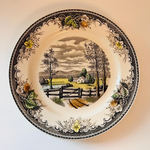 Puede incluir: Un plato de cerámica vintage con una escena de paisaje rural. El plato presenta un borde en blanco y negro con diseños florales y de bayas. La imagen central representa una granja, árboles y una valla en un campo, con un cielo nublado.