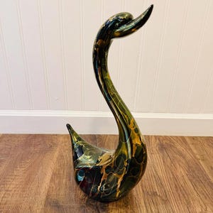 Op de afbeelding: Een decoratief zwaanbeeldje met een lange, gebogen nek. De zwaan is voornamelijk zwart met gouden en groene accenten. Het beeldje is gemaakt van glanzend materiaal en staat op een houten oppervlak.