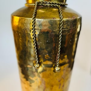 Puede incluir: Un jarrón alto de latón dorado con acabado martillado y un diseño de cuerda alrededor de la parte superior. El jarrón tiene dos borlas decorativas que cuelgan de la cuerda.