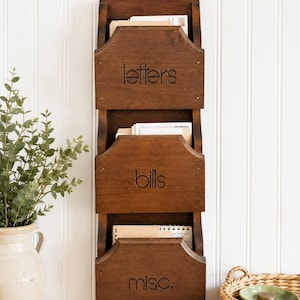 Puede incluir: Un organizador de pared de madera marrón oscuro con tres compartimentos etiquetados como "letters", "bills" y "misc.". El organizador está montado en una pared blanca y tiene un diseño rústico. Tiene un pequeño llavero.