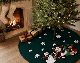May include: A round, dark green Christmas tree skirt featuring white embroidered snowflakes and appliqued figures of a snowman, Santa Claus, and a reindeer. The fabric is solid green with a finished edge, positioned under a lit Christmas tree.