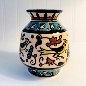 May include: A hand-painted ceramic vase with a cream-colored base, adorned with teal, yellow, and brown bird and floral designs. The vase has a rounded body, a flared rim, and a teal base, showcasing a unique, artistic style.