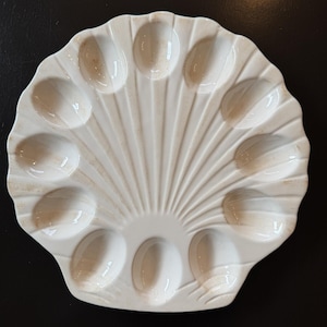 May include: A white ceramic seashell-shaped serving dish with 12 oval indentations. The dish has a ridged, fan-like design and a slightly aged appearance. Suitable for serving appetizers or small portions.