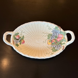 May include: Oval, off-white ceramic serving platter with a woven basket design. The platter features colorful hand-painted grapes, peaches, and leaves. It has two handles and is suitable for serving fruit or other food items.