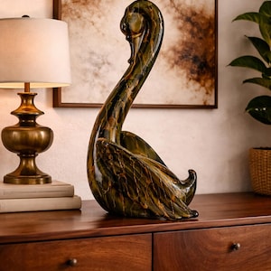 Könnte beinhalten: Eine dekorative Glasskulptur in Form eines Schwans, überwiegend grün mit goldenen Adern, mit langem, gebogenem Hals und ausgebreiteten Flügeln. Der Schwan steht auf einer Holzoberfläche. Ein einzigartiger Dekorationsartikel.