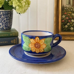 May include: A ceramic teacup and saucer set featuring a vibrant hand-painted yellow sunflower with green leaves against a deep blue background. The cup has a sturdy handle and a white interior, while the matching saucer displays a solid blue finish.