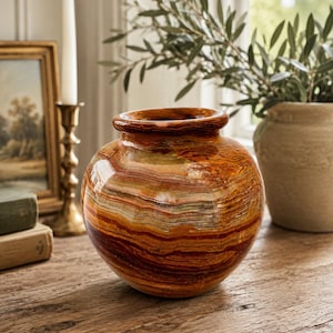 May include: A round, polished stone vase featuring horizontal bands of warm colors including deep rust, burnt orange, cream, and dark brown. The vase has a wide, rounded body with a short, thick neck and a smooth, glossy finish throughout.