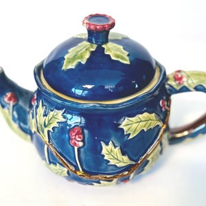 Może przedstawiać: Ciemnoniebieski ceramiczny czajniczek z wypukłym, trójwymiarowym wzorem zielonych liści ostrokrzewu i małych czerwonych jagód. Czajniczek posiada pasującą pokrywkę z falistym uchwytem oraz zakrzywiony uchwyt ozdobiony tym samym motywem roślinnym.