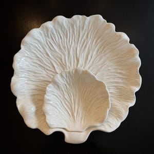 Puede incluir: Un juego de platos de cerámica blanca, con una fuente grande en forma de pétalo con un plato más pequeño a juego anidado en su interior. Los platos tienen una superficie texturizada y veteada, sobre un fondo oscuro.