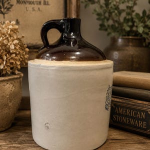 May include: A cylindrical stoneware jug featuring a smooth, off-white body and a dark brown glazed upper section with a single handle. The jug has a narrow neck and a wide base, displaying a simple, functional design typical of traditional pottery.