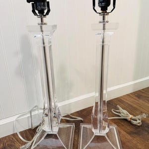 May include: Two clear acrylic table lamps with black lamp holders and silver stems. Each lamp has a square base and a tall, rectangular body. The lamps are plugged in and ready to use.