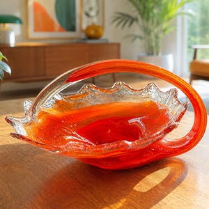 May include: A vibrant orange and clear glass basket with a ruffled, textured rim and a curved handle. The bowl features a deep orange centre that transitions into clear glass edges, showcasing a bubbled texture throughout the entire piece.