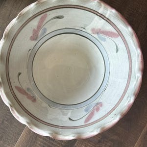 May include: A large, off-white ceramic bowl with a scalloped edge. The bowl features a series of concentric rings in gray and pink, with stylized floral accents in pink and blue. The bowl is likely handmade.