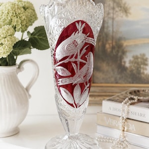 Könnte beinhalten: Eine klare Kristallvase mit gewelltem Rand und Rautenmuster. In der Mitte befindet sich eine markante ovale Einlage aus rotem Glas mit einer Gravur von zwei Vögeln, die auf Zweigen zwischen Blättern sitzen. Der Fuß ist kreisförmig und facettiert.