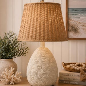 Puede incluir: Lámpara de mesa beige con pantalla tejida y base decorada con un patrón de conchas marinas. La lámpara está sobre una mesa de madera, con un jarrón, coral y libros cerca. La lámpara mide aproximadamente 61 cm de altura.