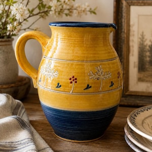 May include: A ceramic pitcher featuring a glossy yellow upper body and a deep blue base. The middle section is decorated with embossed white tree-like motifs and small red dots. It has a curved handle and a blue rim, showcasing a handcrafted aesthetic.