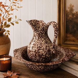 May include: A ceramic pitcher and matching basin set featuring a brown and cream speckled pattern. The pitcher has a curved handle and a scalloped rim, while the basin has a wide, shallow bowl shape with a matching scalloped edge design.