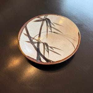 May include: A round ceramic plate with a bamboo design. The plate has a cream-colored center with a brown rim. The bamboo is depicted in black and brown, with detailed leaves and stalks. The plate is likely used for serving food.