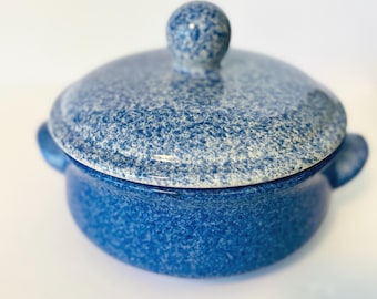 May include: A blue speckled ceramic casserole dish with a matching lid. The dish has two small handles and a round knob on top of the lid. The speckled pattern is a mix of dark and light blue.